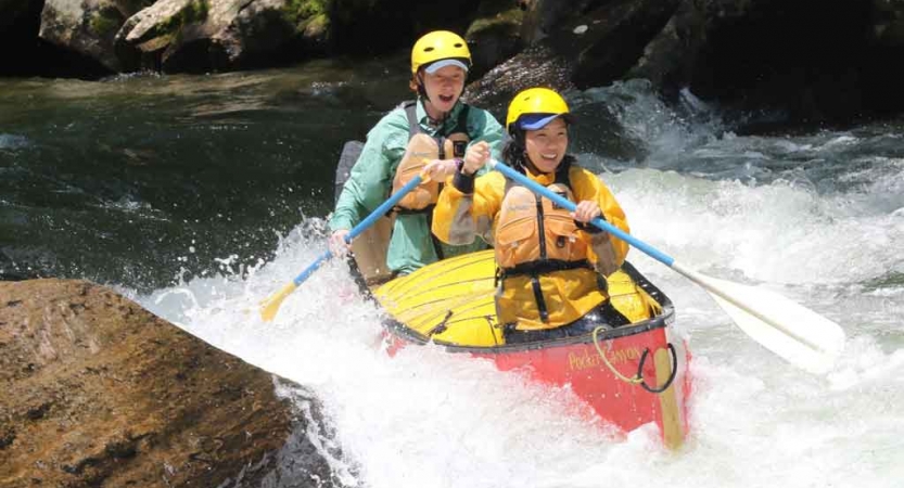 two students wearing safety gear paddle a canoe navigate whitewater in north carolina on an outward bound trip